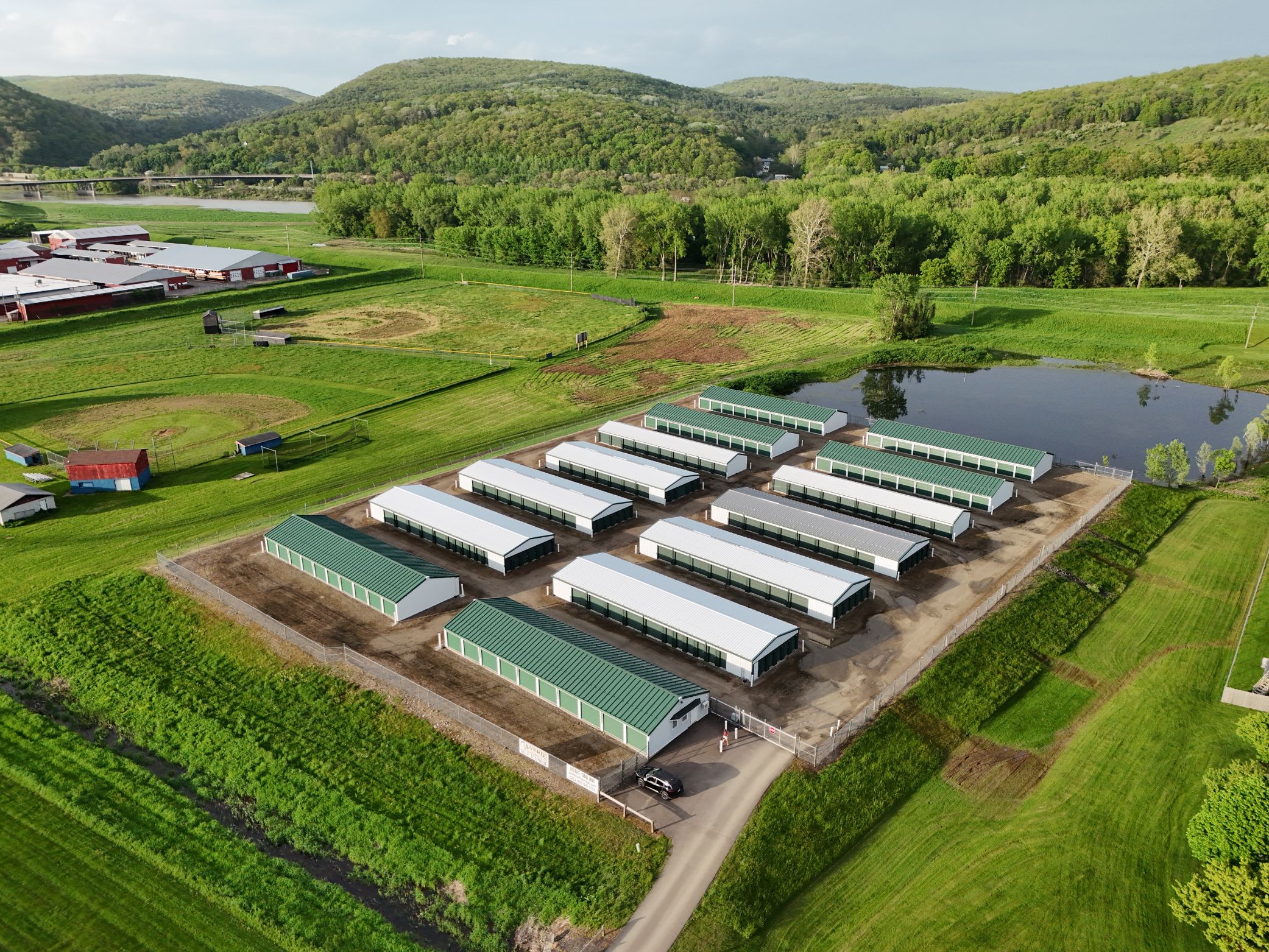 Aerial view of A-Verdi Self Storage facility with green and white storage buildings, gated entry, pond, and wooded hills in the background.
